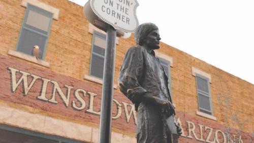 Statue of a man standing beside a sign that reads "Standin' on the Corner" in Winslow, Arizona, with a historic building in the background.