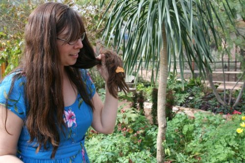 A woman with long hair in a blue dress stands in a lush garden, playfully touching her hair.