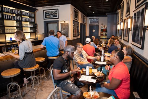 A lively bar scene with patrons enjoying drinks and food, seated at tables and the bar, in a cozy, stylish setting.