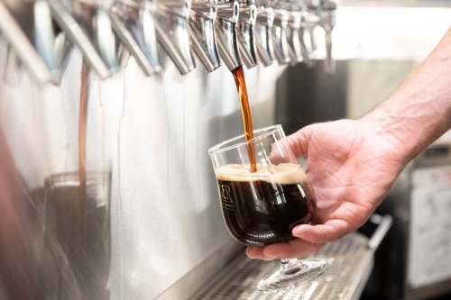 A hand pours dark beer from a tap into a glass, with multiple taps visible in the background.