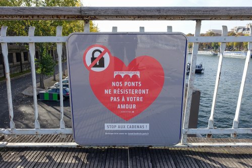 Sign on a bridge urging against placing love locks, featuring a heart and a message in French about love and bridges.