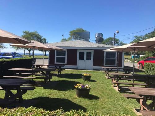 A small red building with a gray roof, surrounded by picnic tables and umbrellas, set in a grassy area near water.