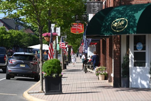 A charming street scene with shops, flags, and people walking on a sunny day. Greenery and planters line the sidewalk.