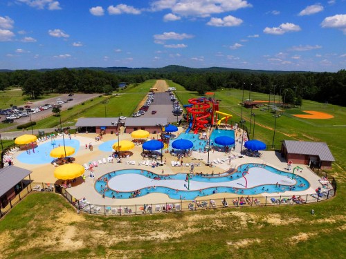 Aerial view of a vibrant water park with pools, slides, and colorful umbrellas, surrounded by green fields and a parking area.