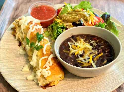 A plate with a rolled enchilada topped with sauce, black beans, salad, guacamole, and salsa on the side.