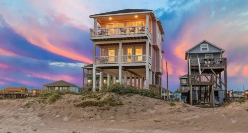 A beach house with multiple balconies stands against a colorful sunset sky, surrounded by sandy dunes.