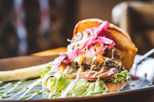 A close-up of a juicy burger topped with lettuce, cheese, pickled onions, and drizzled with sauce, served on a plate.