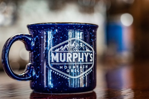 A blue ceramic mug with "Murphy's Mountain Grill" and mountain graphics, set on a wooden table.