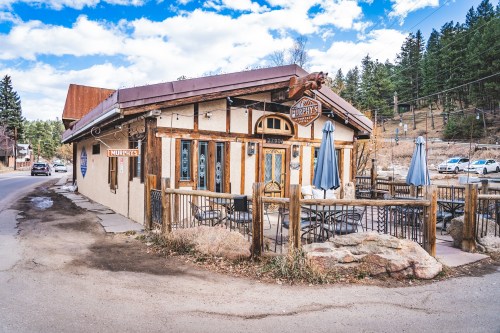 A charming rustic restaurant with outdoor seating, surrounded by trees and a clear blue sky.