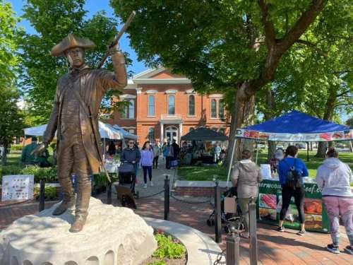 A bronze statue of a soldier stands in a park, with a farmers market and people browsing in the background.