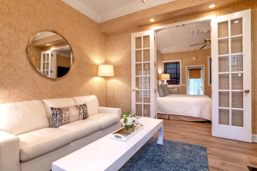 Cozy living area with a white sofa, coffee table, and a view of a bedroom through French doors. Warm, inviting decor.