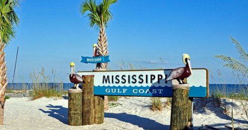 Welcome sign for Mississippi Gulf Coast, featuring palm trees and pelican sculptures against a blue sky and sandy beach.
