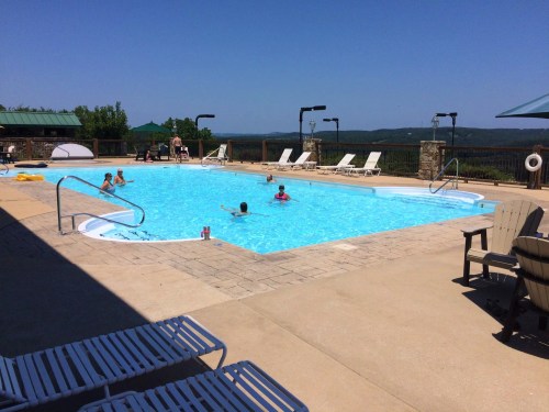 A sunny pool area with people swimming, lounge chairs, and scenic views in the background.