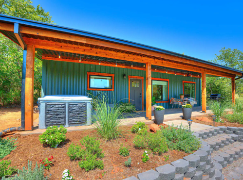 A modern blue house with a wooden porch, hot tub, and landscaped garden under a clear blue sky.