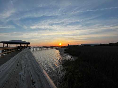 Sunset over a calm waterway, with a wooden pier extending into the distance and silhouettes of people enjoying the view.