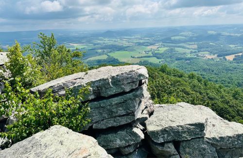 A rocky outcrop overlooks a lush green valley and rolling hills under a partly cloudy sky.