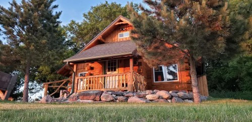 A cozy wooden cabin surrounded by trees and rocks, with a porch and warm sunlight illuminating the scene.