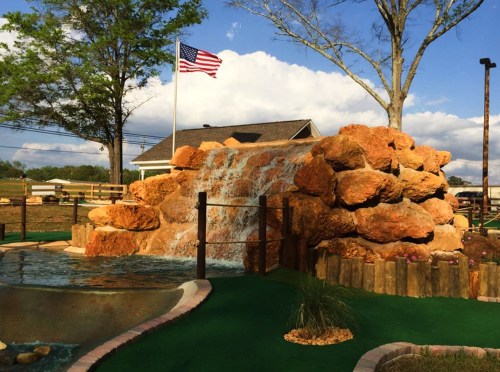 A small waterfall cascades over rocks near a pond, with a flag flying in the background and trees lining the area.