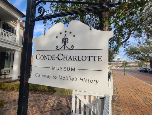 Sign for the Condé-Charlotte Museum, stating "Gateway to Mobile's History," with trees and buildings in the background.