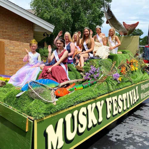 A colorful parade float decorated with grass and flowers, featuring young women in dresses waving to the crowd.