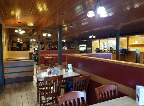 Cozy restaurant interior with wooden tables, chairs, and warm lighting, featuring a counter and kitchen area in the background.