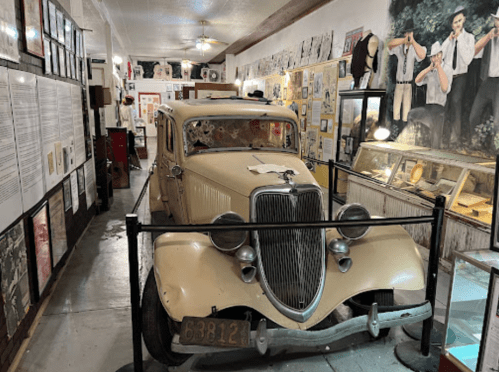 A vintage car displayed in a museum, surrounded by historical artifacts and photographs on the walls.