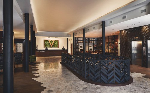 Modern hotel lobby featuring a reception desk, stylish decor, and a unique patterned railing. Warm lighting enhances the ambiance.
