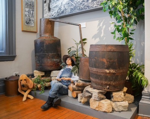 A vintage display featuring a mannequin in overalls, a dog statue, and old barrels against a wall with historical photos.