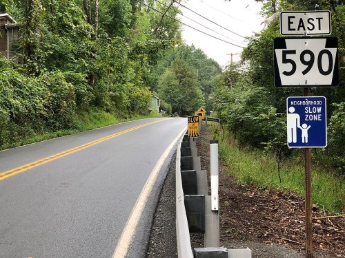 A road sign for East 590, with a "SLOW" warning and a neighborhood slow zone sign, surrounded by greenery.