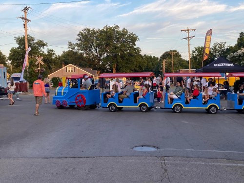 A colorful train ride with children and adults, surrounded by a lively outdoor event with food stalls and trees.