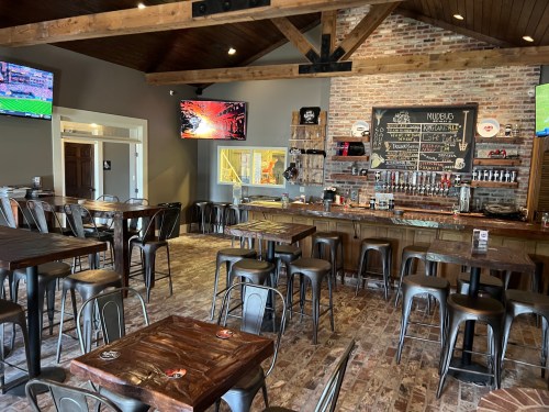 Interior of a bar with wooden tables, metal chairs, a brick wall, and multiple TVs displaying sports.