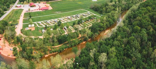 Aerial view of a campsite along a river, surrounded by greenery and open fields, with parked vehicles and tents visible.