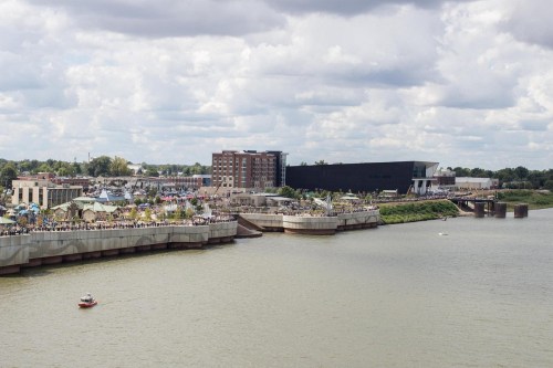 A riverfront scene with a crowd, buildings, and a boat on the water under a partly cloudy sky.