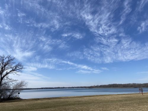 A serene lake scene with a clear blue sky and wispy clouds, framed by grass and a few trees on the shore.