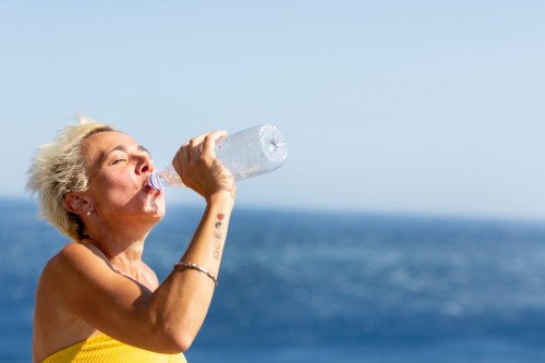 A woman with short blonde hair drinks water from a bottle against a backdrop of the ocean and clear blue sky.