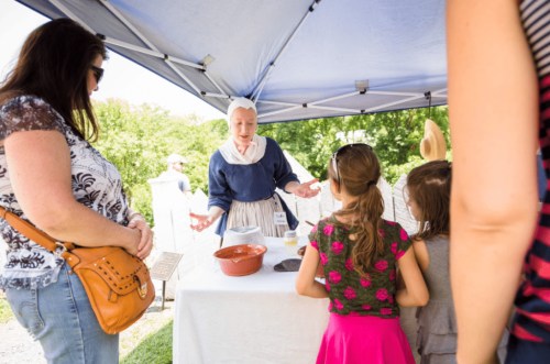 A historical reenactor in period clothing demonstrates cooking to a small group under a tent.