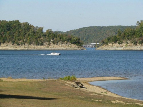 A serene lake scene with a boat cruising through calm waters, surrounded by green hills and sandy shores.