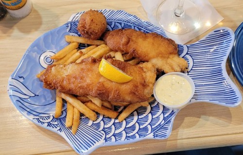 A blue fish-shaped plate with two pieces of fried fish, fries, a lemon wedge, a hushpuppy, and a side of tartar sauce.