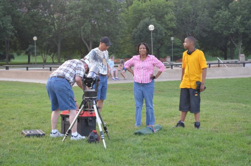 A group of four people set up a telescope in a park, with trees and a playground in the background.