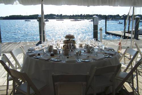A beautifully set round table with white chairs, overlooking a sparkling waterfront and boats in the distance.