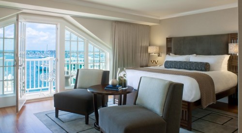 A stylish hotel room with a king bed, two chairs, and a view of the ocean through open balcony doors.