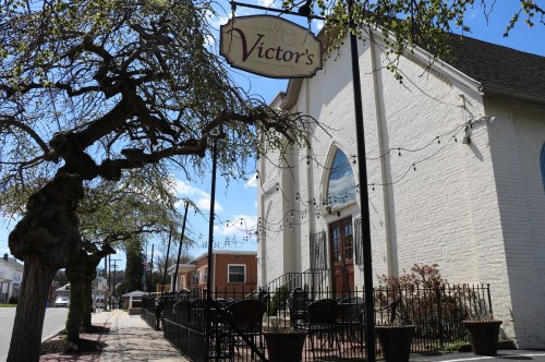 A white building with a sign reading "Victor's," surrounded by trees and outdoor seating on a sunny day.