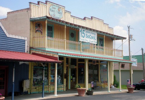 Colorful two-story building with a sign celebrating 15 years, featuring a porch and various storefronts.