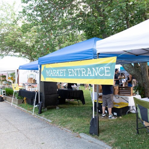 A colorful sign reading "Market Entrance" points left, with vendor tents and greenery in the background.