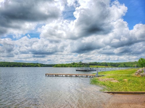 A serene lake scene with a wooden dock, lush greenery, and dramatic clouds in a blue sky.