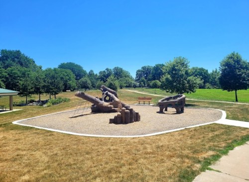 A playground with unique wooden structures surrounded by grass and trees under a clear blue sky.