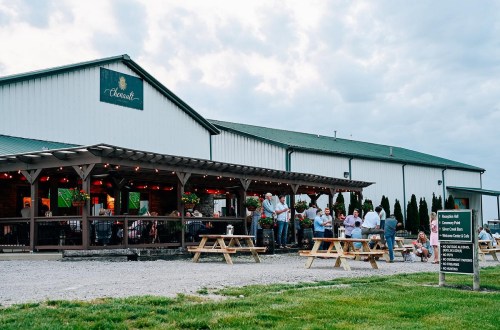 A rustic restaurant with outdoor seating, people dining, and a cloudy sky in the background. Greenery surrounds the area.