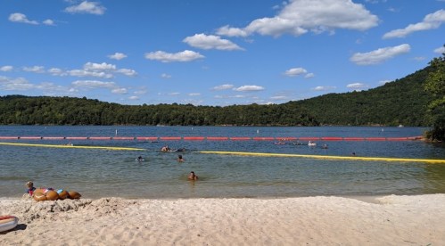 A sandy beach by a lake with swimmers, surrounded by green hills and a blue sky with fluffy clouds.