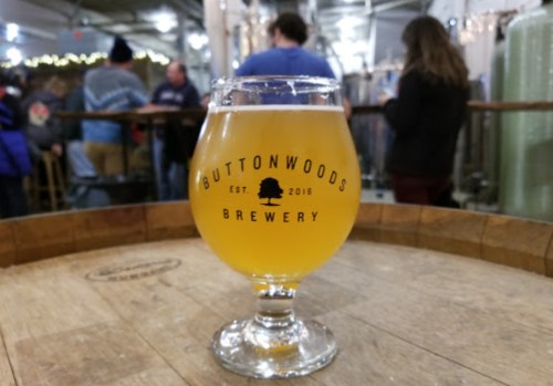 A glass of beer from Buttonwoods Brewery sits on a wooden barrel, with people socializing in the background.