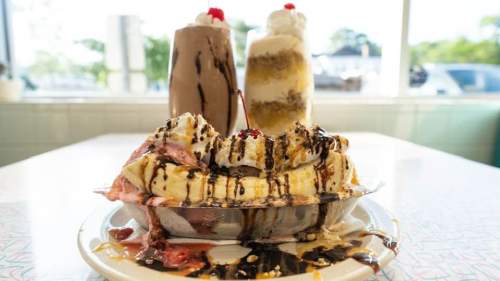 A colorful banana split topped with whipped cream, chocolate syrup, and a cherry, with milkshakes in the background.